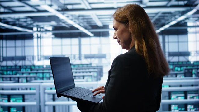 System administrator in server hub checking recovery plan on laptop, monitoring energy consumption. IT expert uses notebook in data center workspace, ensuring rigs are functioning optimally, camera B