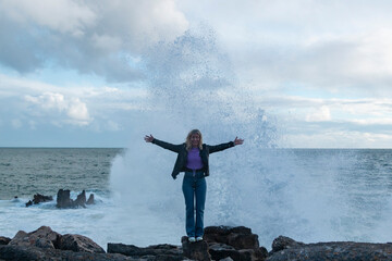 Blonde woman with the sea in the background and a wave breaking behind her during sunset