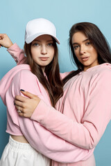 Two Young Brunette Woman Models Posing on a Blue Background in Pink Clothes, White Cap, Sunglasses. Beautiful Smiles, Lovely Girls Friends.