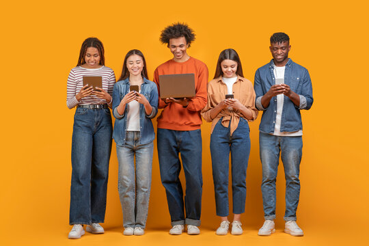 This image shows a group of five young adults standing in a row against a yellow background. They are all looking down at their devices, which include a tablet, a smartphone, and a laptop