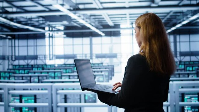 System administrator in server farm using laptop to patch network issues leading to slow data transfer rates. IT support staff member in data center fixing errors affecting supercomputers, camera B