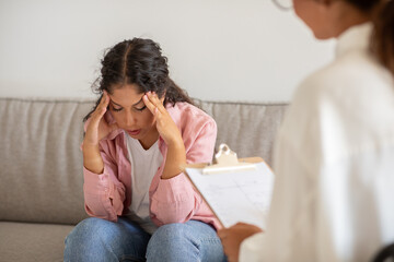 This image shows a woman sitting on a couch, holding her head in her hands, looking distressed. She...