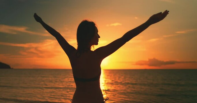 A woman is standing on Koh Ma Beach in Thailands Koh Phangan Island as the sun sets behind her, casting a golden glow over the sandy shore and calm ocean waves.