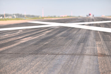 A white cross on an airport runway prohibits landing of airliners due to repairs. Close-up view of the texture of aircraft landing gear tire tracks. Airport structures and an airplane are blurred