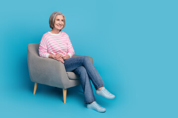 Full size photo of attractive retired woman sit armchair look empty space dressed stylish striped clothes isolated on blue color background