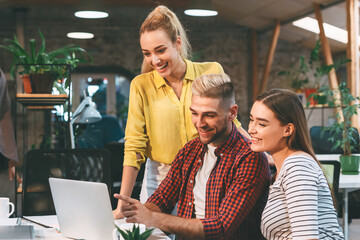 Three young colleagues, two women and a man, smile and discuss work while gathered around a laptop in a well-lit, modern office space.