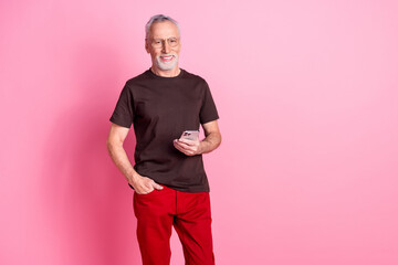 Portrait of good mood senior man with beard dressed t-shirt in glasses hold smartphone arm in pocket isolated on pink color background