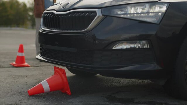 Close-up shot of front end of black car running over orange traffic cone while performing exercise at lesson, driving instructor is standing nearby