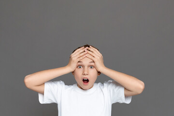 Teenage boy with an expression of sheer surprise, hands on head, isolated on a grey background,...