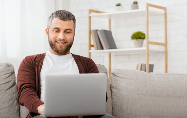 A man is seated comfortably on a couch, engrossed in his laptop screen. He appears focused as he...