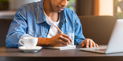 Cropped of guy is seated at a table with a laptop open in front of him. He is holding a cup of coffee, appearing focused on his work or task at hand in a casual setting.