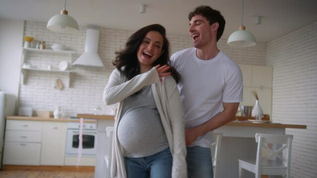 Happy funny active carefree couple people pregnant family love Caucasian man guy male boyfriend husband having fun fooling pregnancy belly wife girlfriend woman girl female dancing home kitchen dance