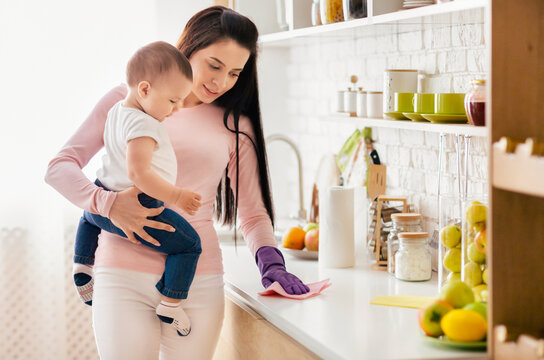 A mother is cleaning the kitchen counter with a cloth while holding her baby. The kitchen has a bright, organized, and clean appearance. Fruit and kitchen utensils are visible on the counter - Powered by Adobe