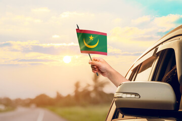 Waving the Mauritania flag against the sunrise or sunset from a car driving along a country road. Holding the Mauritania flag, traveling by car, on a weekend trip.
