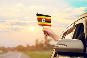 Waving the Uganda flag against the sunrise or sunset from a car driving along a country road. Holding the Uganda flag, traveling by car, on a weekend trip.