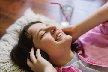 A teengirl is lying on her back on a wooden floor, wearing headphones and a pink shirt. She has her eyes closed and is smiling, seemingly relaxed and listening to music.