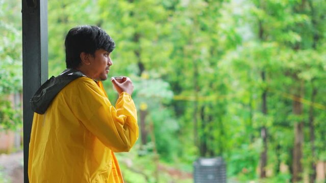 Rear view shot of an Indian man wearing yellow rain jacket drinking tea while looking at the rain fall during the monsoon season in the middle of the forest at Saputara in Gujarat, India.