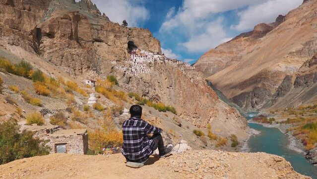 Rear view shot of an Indian male tourist sitting in front of the Phugtal Monastery at Zanskar Valley in Ladakh, India. Man looks at the 2500 year old monastery built in a cave. Travel background.