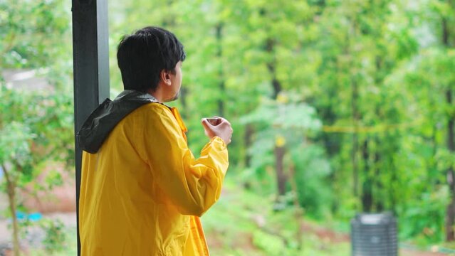 Rear view shot of an Indian man wearing yellow rain jacket drinking tea while looking at the rain fall during the monsoon season in the middle of the forest at Saputara in Gujarat, India.