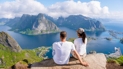 A couple sits on a mountaintop overlooking a stunning view of the Lofoten Islands in Norway, with...