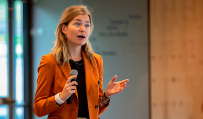 Woman in orange blazer speaking into a microphone during a presentation. Business and public speaking concept. Design for poster, banner, print