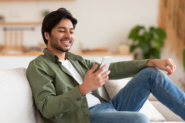A man is sitting on a couch in a home setting, smiling while looking at his smartphone. He is wearing a green shirt, white t-shirt, and blue jeans.