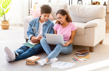 African American teen couple sitting on the floor in their home, studying together. The girl is using a laptop while the guy is taking notes in a notebook.