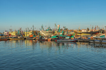 A panoramic view of a harbor filled with docked fishing boats, with the setting sun casting a warm glow on the water and boats.