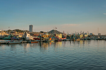 A panoramic view of a harbor filled with docked fishing boats, with the setting sun casting a warm glow on the water and boats.