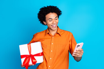 Photo of cheerful positive guy dressed orange shirt holding present box chatting device isolated...