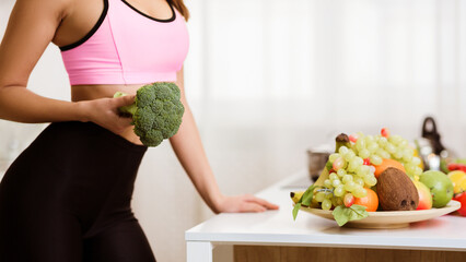 A woman wearing a pink sports bra and black leggings, holds a large head of broccoli in front of a...