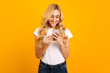 A young woman with long blonde hair, wearing a white t-shirt and blue jeans, is smiling and looking at her smartphone in front of a bright yellow background. She appears happy and engaged