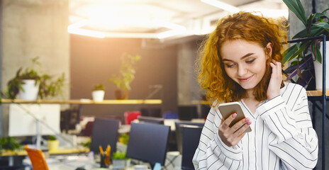 A woman is sitting in an office, focused on her phone screen, reading or typing a message. The office desk is cluttered with papers and a computer monitor is visible in the background.