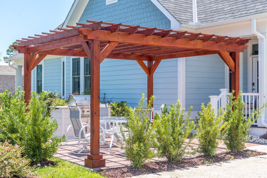 Trendy outdoor patio pergola shade structure made of dark wood, awning and patio roof, white dining table, white chairs, metal grill surrounded by landscaping in a luxury American neighborhood