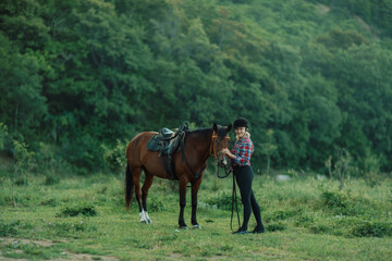 Happy blonde with horse in forest. Woman and a horse walking through the field during the day. Dressed in a plaid shirt and black leggings.