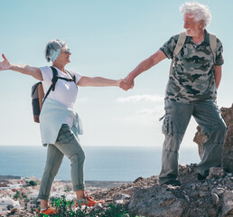 Happy active senior couple in outdoor excursion hiking in mountain with backpack  enjoying healthy lifestyle. Bearded man helping woman climb up. Horizon over water on background