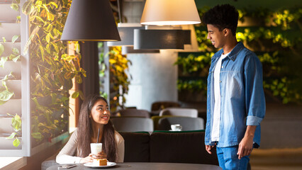 Two African American friends are enjoying a casual meeting in a cozy cafe in the evening. One is...