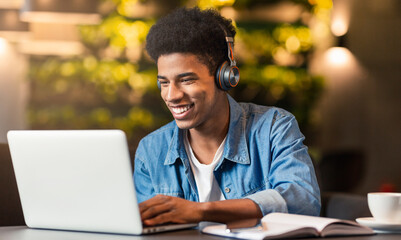 A young black guy sits at a table in a coffee shop, wearing headphones and focusing intently on his...