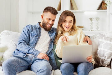 A man and woman are seated on a couch, both focused on a laptop screen in front of them.