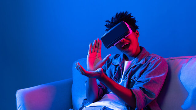 Young African American looking VR enjoy watching fantastic 3D pointing interesting object technology blue neon light virtual reality metaverse new world at living room sitting on sofa. Contrivance.