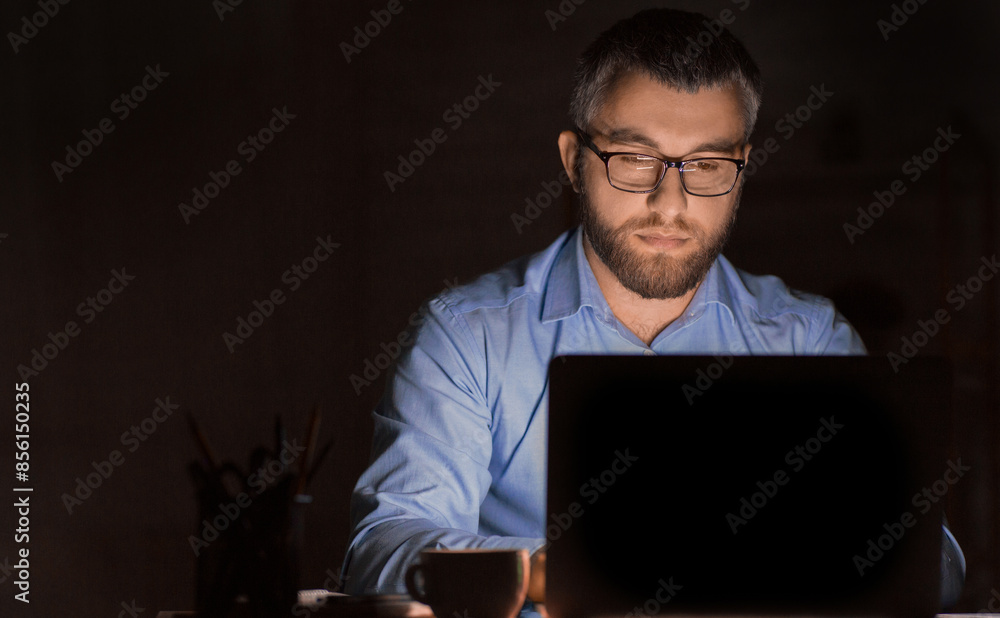 Poster a man wearing glasses is intensely focused while working on his laptop in a dimly lit office environ