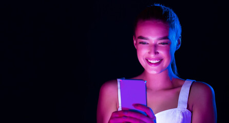 A young woman is sitting alone in a dark room, illuminated only by the glow of her phone screen, engrossed in her device, scrolling and typing. The light from the phone casts shadows on her face