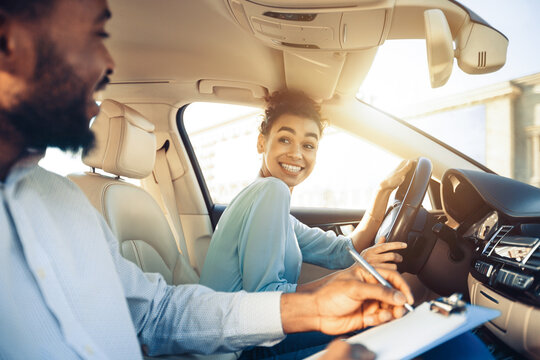 A driving instructor sits in the passenger seat of a car while a student driver sits in the drivers seat and turns to look at the instructor. The instructor is using a pen to write on a clipboard.