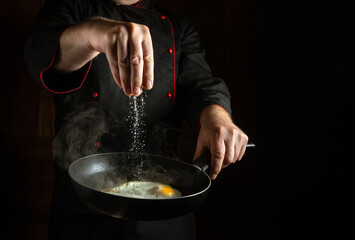 Fototapeta premium Chef salting fried egg by hand in frying pan. Concept of cooking egg breakfast in dark kitchen.