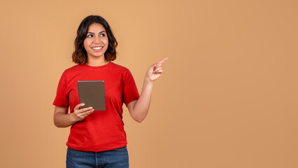 A young middle eastern woman, wearing a red t-shirt and blue jeans, is holding a tablet and pointing her right hand to copy space while smiling. She is standing against a light brown background.