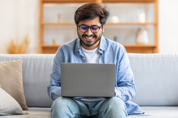 Indian man seated on a couch indoors, engrossed in using a laptop computer for work or leisure...