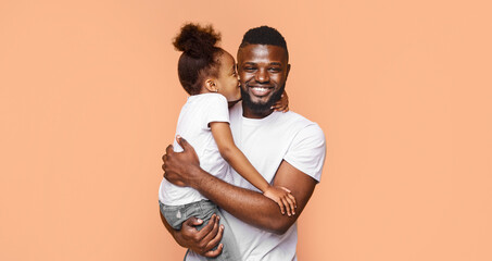Obraz premium A happy Black father is holding his little daughter and smiling at the camera. They are standing against a peach-colored background