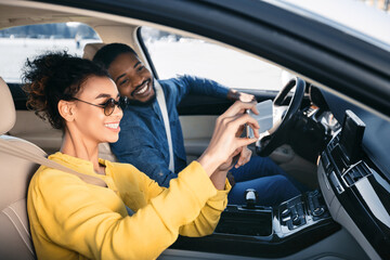 African American woman in sunglasses sits in the passenger seat of a car and takes a selfie with her boyfriend who is driving, both of them smiling at the camera.