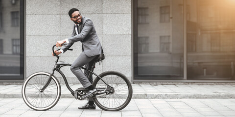 Fototapeta premium An African American businessman is biking to the office and standing by a grey wall in an urban area. The scene is modern and stylish, emphasizing a blend of professional and active lifestyle.