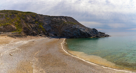 Vista panorámica de la playa Piccola en Argentiera, Cerdeña, Italia. La playa, con arena dorada y guijarros, está rodeada de imponentes acantilados y colinas verdes. El mar esmeralda es sereno y claro